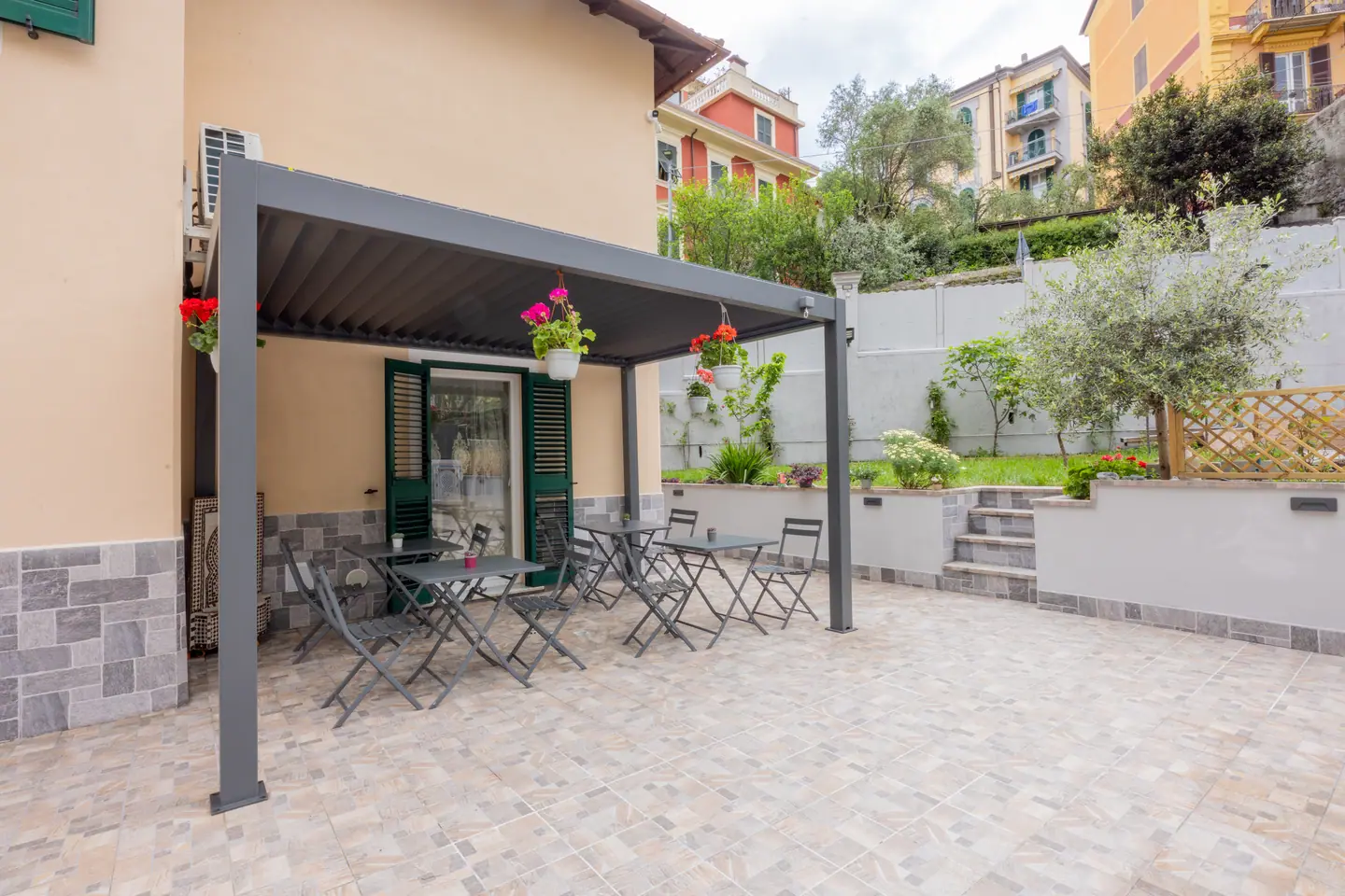 Il cortile sul retro: pergola in alluminio, tavolini da caffè, gerani appesi, ulivo e le case pastello della collina alle spalle