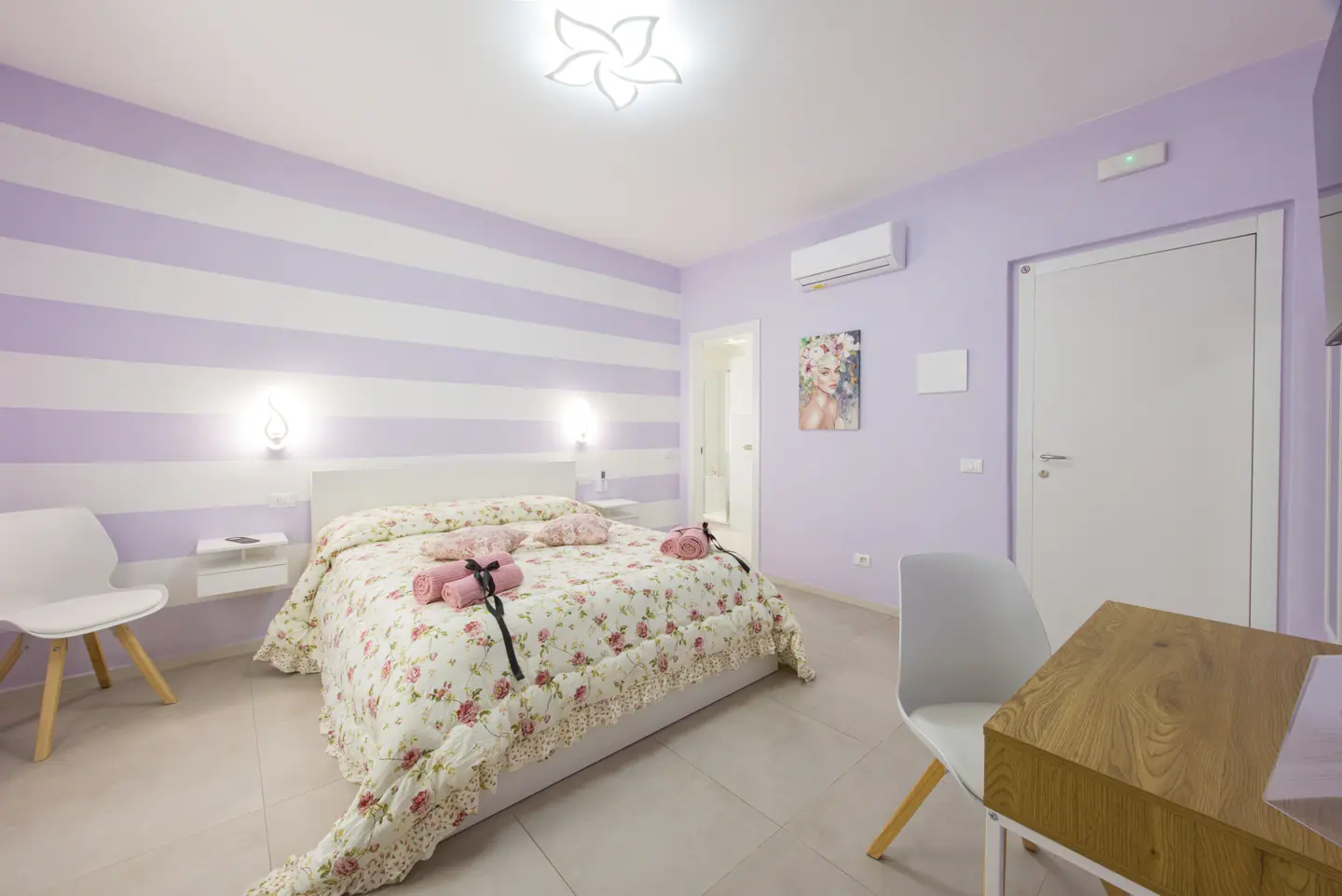 La camera Tellaro: letto matrimoniale con trapunta a rose, parete a righe lilla e bianche, applique accese